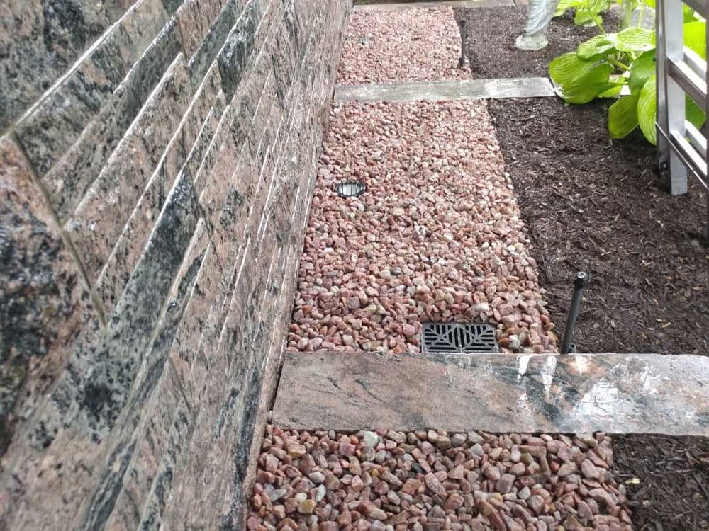 Landscape with granite wall, pink gravel pathway, and drainage features in garden setting.