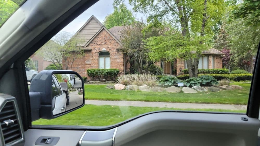Brick house surrounded by greenery and landscaping visible from a car window.