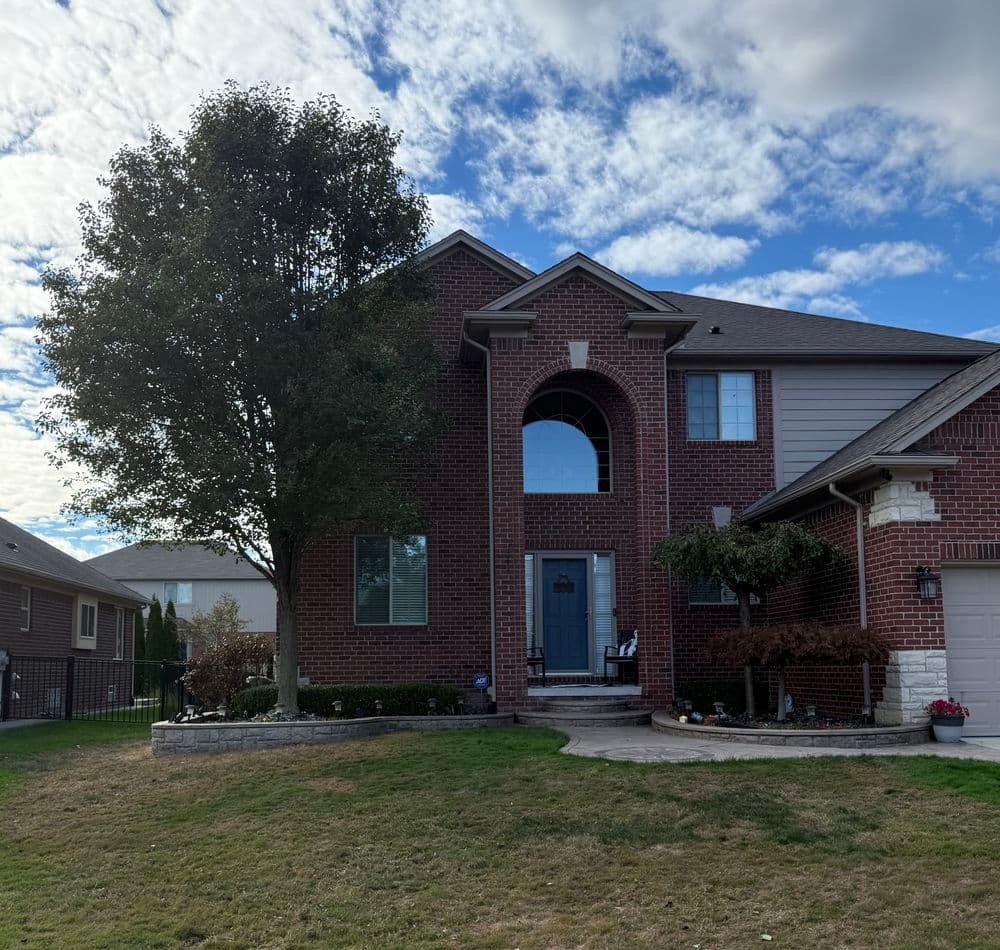 Two-story red brick house with large tree and blue front door, set under a cloudy sky.
