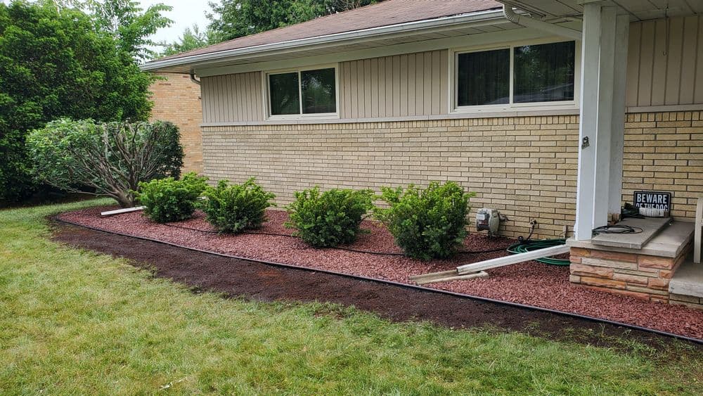 Landscaped home exterior with shrubs, red mulch, and garden hose near brick foundation.