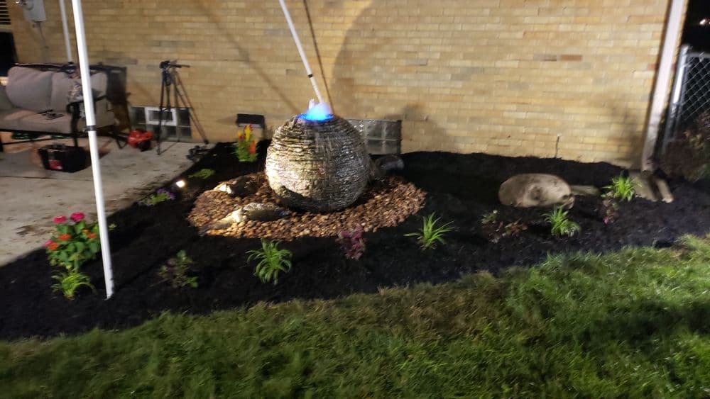 Outdoor garden with a stone fountain, decorative plants, and mulch at night.