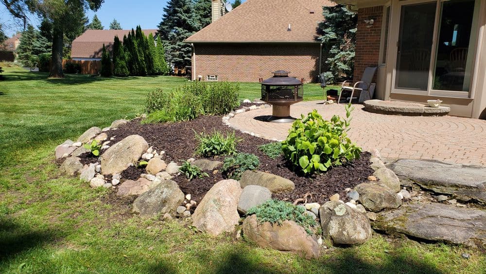 Landscaped garden with rocks, plants, and a decorative fire pit in a sunny backyard setting.