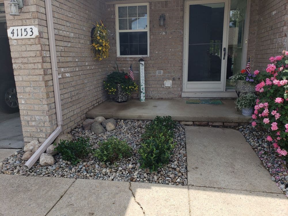 Front porch with brick wall, flower pots, and landscaped rock garden featuring greenery and flags.