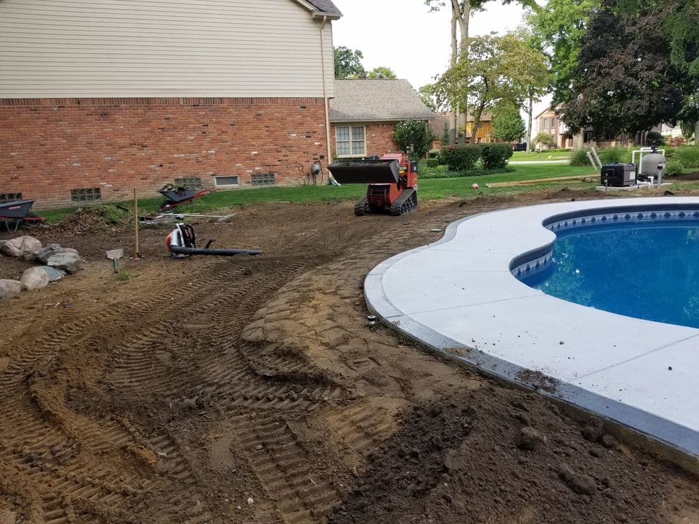 Construction equipment near a pool in a landscaped yard with freshly tilled soil.