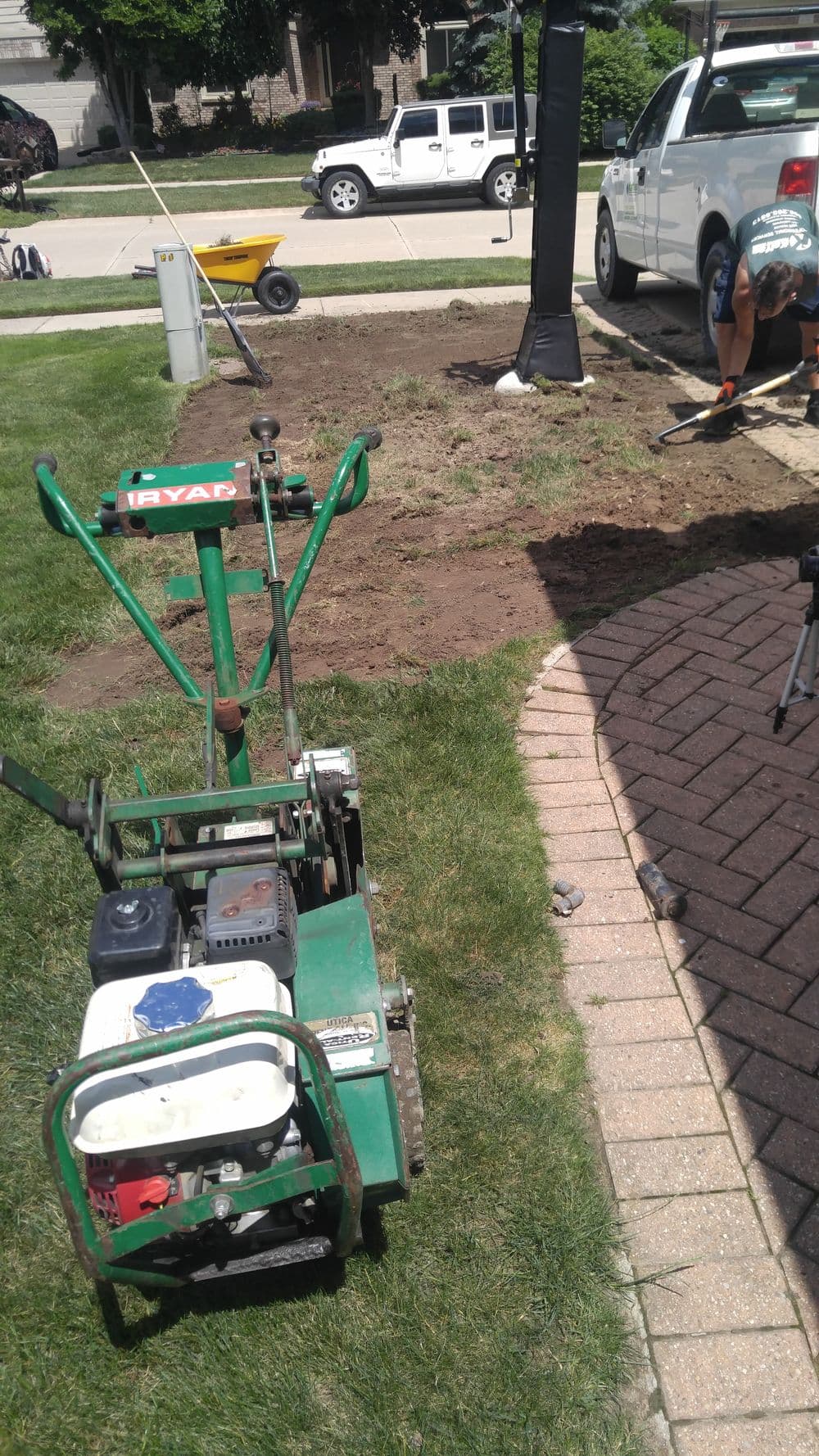 Lawn aerator on grass with parked vehicles and landscaping tools nearby.