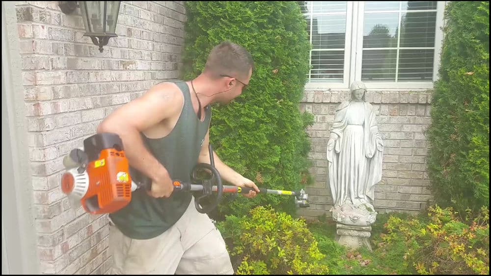 Man using a string trimmer near a garden statue in a well-maintained yard.