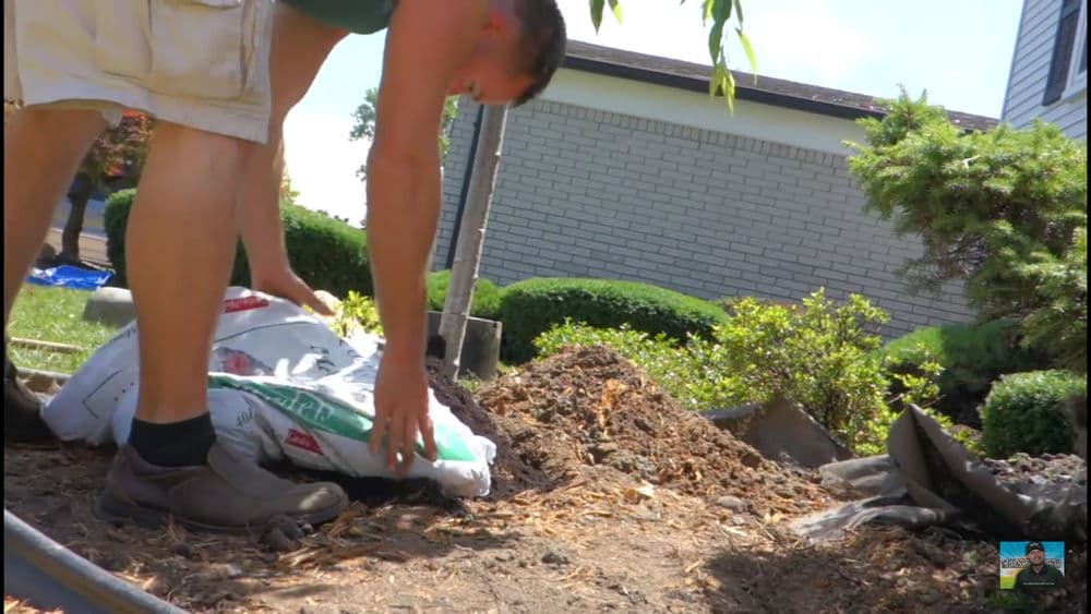 Man in shorts lifting a bag of mulch in a landscaped garden setting.